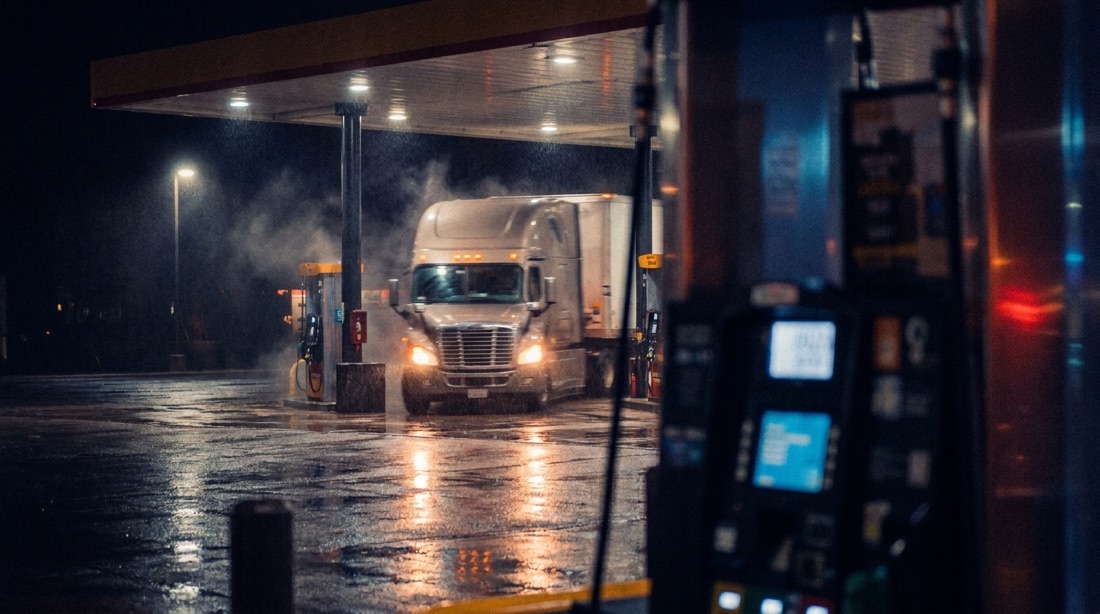 Fuel island at a truck stop at night, pump numbers glowing over wet pavement