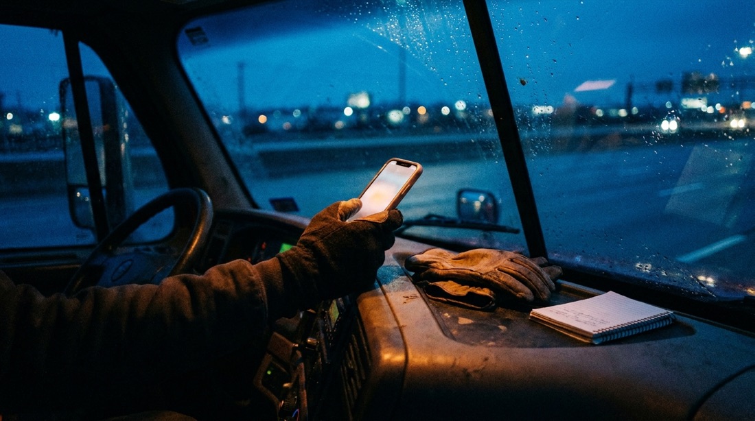 Truck driver checking a phone above the dashboard at night, notebook and gloves on the dash